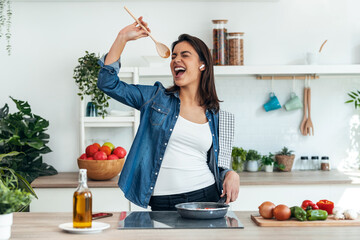 Beautiful young woman cooking while singing and listening music with earphones in the kitchen at home.