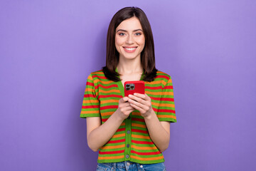 Portrait of cheerful positive girl with stylish hairdo dressed striped cardigan typing email on smartphone isolated on violet background