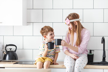 Happy children holding black cups sitting in modern cozy kitchen enjoying morning tea or coffee. Siblings spending weekend together at home. Good morning, start new day concept.
