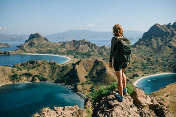 A young girl with a backpack stands on top of a mountain and admires the panorama of Padar Island in Indonesia, rear view. - obrazy, fototapety, plakaty