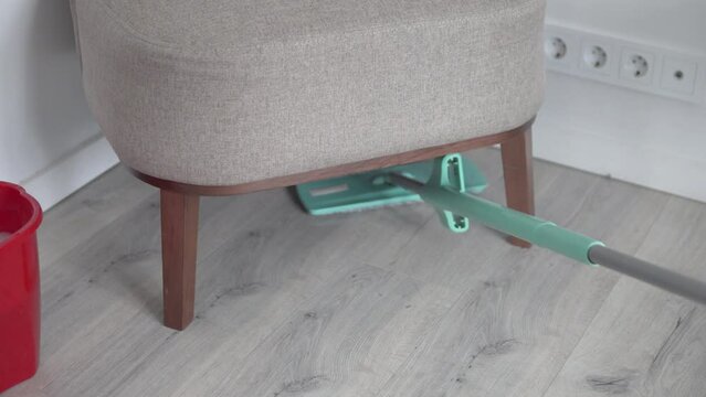 A man mops the floors under a chair on a leg in an apartment