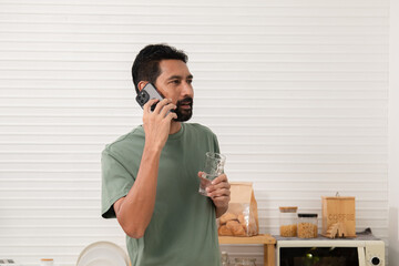 Happy young Asian man using a smartphone drinking clean water in the morning immediately after waking up. Healthy smart man cleanses his body by drinking water after waking up first.