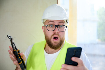 Engineer with white helmet using his smart phone at the construction site. Portrait of construction worker in uniform playing games or makes bets on mobile phone during break. Fast delivery. Win.