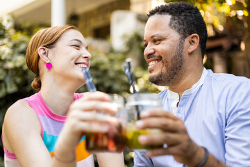 A multiracial couple have fun drinking mojitos happily in an outdoor venue. The African man and the Caucasian woman toast with cocktails while looking at each other. Concept of summer celebration.