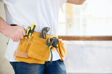 Cropped view of the man in white T-shirt and blue jeans with yellow tool belt near concrete or cement wall and wide light window.