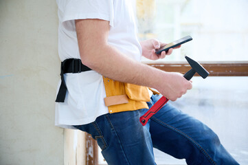 Crop view of engineer with orange tool belt using his smart phone at the construction site. Crop portrait of mature construction worker in uniform browsing social media on mobile phone during break.