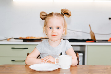 Portrait of a caucasian girl sitting at a table with an empty plate and a mug in the kitchen, a hungry child waiting for food indoors