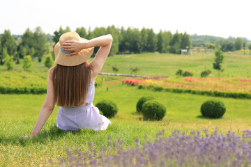 Obraz premium A young girl in a dress sits on green grass in a natural park on a summer day. Spend time alone in nature. Calm atmosphere. Back view. A girl in a summer hat looks at the landscape