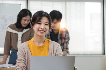 A focused young Asian female marketing assistant is working on her work on her laptop at her desk while her coworkers are working in the room.