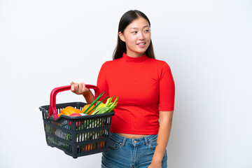 Fototapeta na wymiar Young Asian woman holding a shopping basket full of food isolated on white background looking to the side and smiling