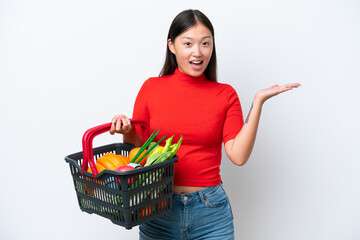 Obraz na płótnie Canvas Young Asian woman holding a shopping basket full of food isolated on white background with shocked facial expression