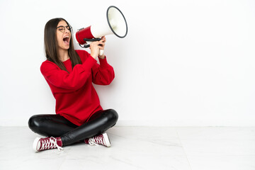 Young woman sitting on the floor isolated on white background shouting through a megaphone