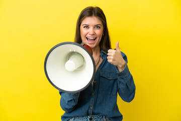 Young caucasian woman isolated on yellow background shouting through a megaphone to announce something and with thumb up
