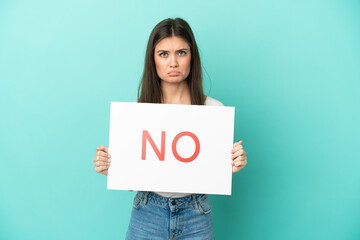 Young caucasian woman isolated on blue background holding a placard with text NO with sad expression