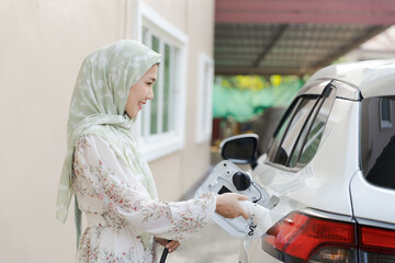 Muslim Islamic women using a charging pump into an EV car electric vehicle, traveling to places using automobile transportation, charging at home, eco environmental friendly green energy technology.