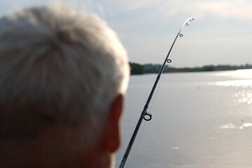 Old man fishing. Senior gray haired fisherman throws a spinning from shoreside at sunset, twisting a coil. Positive elderly male angling at lake, rotating reel rod. Retired fisher enjoying his weekend