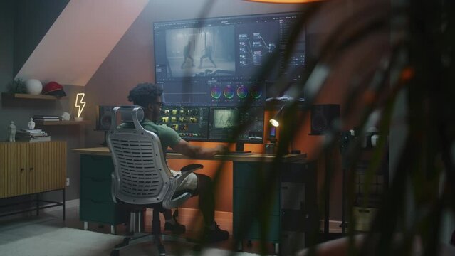 African American male editor makes color grading, works at home office. Action movie footage and program interface on multi-monitor computer and big digital screen. Concept of film post production.