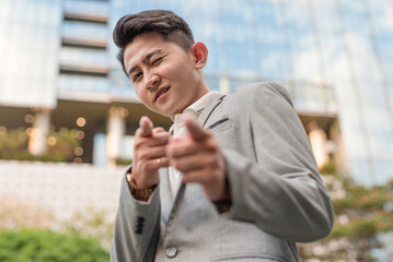 A young confident corporate asian man in his early 20s winks and makes a finger gun gesture just outside the city.