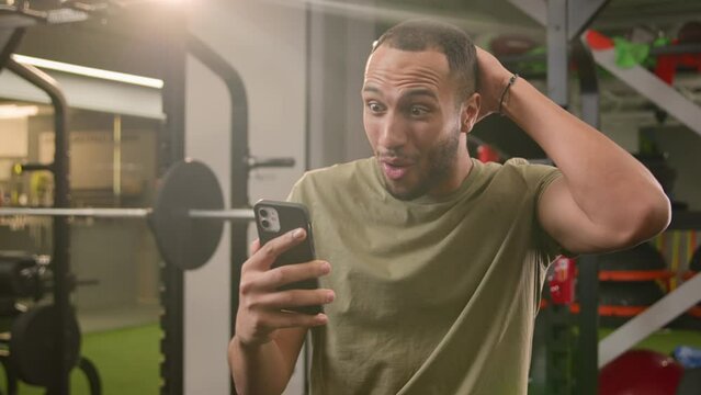 Shocked wonder surprised excited African American man trainer guy sportsman male athlete bodybuilder in sport club gym happy win victory achieve success with mobile phone message winning say wow shock