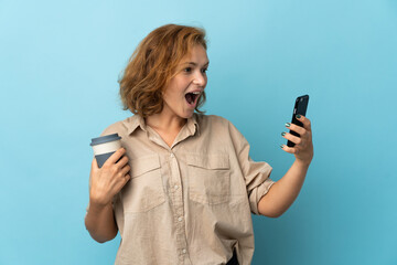 Young Georgian woman isolated on blue background holding coffee to take away and a mobile