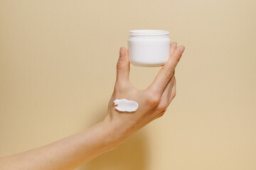 Female hand with smear holding white mockup jar of facial moisturizer on beige isolated background. The concept of beauty treatment, skin care and spa