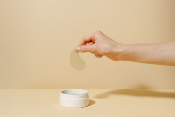 Female hand holding sample of green algae extract eye patch over white jar of product on beige isolated background. The concept of natural product for moisturizing, from dark circles under the eyes