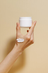 Vertical shot of a female hand with a smear of moisturizer holding a jar of white cream mockup on a beige isolated background. Concept of  face care, moisturizing, and sun protection