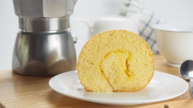 Coffee time with a portion of sponge cake roll filled with pineapple jam in white ceramic dish and a cup of coffee from moka pot coffee maker on wooden table.