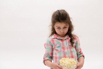 Lovely baby girl in stylish pink and blue plaid shirt, holding bowl with popcorn, isolated on white background. People. Children. Delicious fast food. Entertainment. Leisure. Lifestyle. Cinema concept