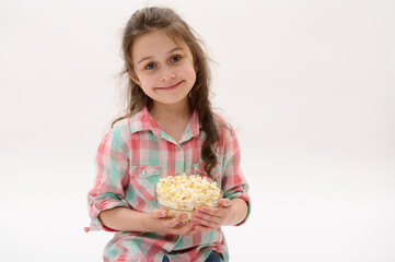 Caucasian adorable little child girl, first grader, primary school student, in stylish checkered shirt, smiling broadly looking at camera, posing with bowl of sweet popcorn on white studio background