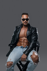 Fototapeta premium Attractive male model with a muscular shirtless torso poses in ripped jeans, a black leather jacket, and dark sunglasses while seated on a studio chair against a grey backdrop