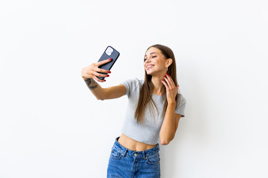 Young woman laughing and showing peace sign while taking selfie photo on cellphone isolated over white background