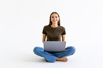 Portrait of a smiling casual woman holding laptop computer while sitting on a floor isolated over...