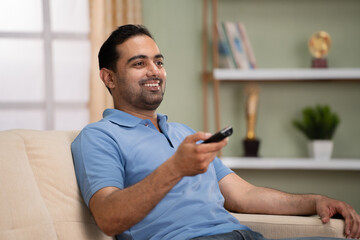 Relaxed Happy indian man watching live sports match on tv or television while sitting on sofa at...