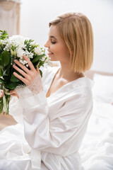 Obraz na płótnie Canvas happiness, cheerful bride with blonde hair sitting on bed and smelling white flowers, bridal bouquet, young woman in white robe, beautiful, excitement, feminine, blissful, wedding preparation
