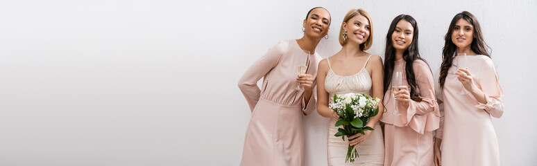 positivity, happy bride in wedding dress holding bridal bouquet and standing near interracial bridesmaids on grey background, champagne glasses, racial diversity, fashion, banner