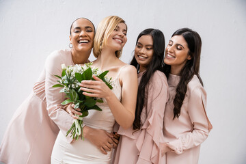 excitement, multicultural bridesmaids hugging happy bride in wedding dress with bridal bouquet,...