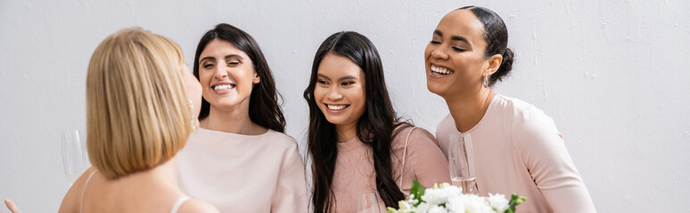 wedding preparations, cheerful interracial bridesmaids looking at blonde bride with bouquet on grey...