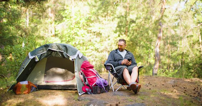 Breath of fredom in a morning forest - runaway from noise of big city. Middle aged man relaxing in a travel armchair in a forest next his tent. Rest in nature, as a guarantee of a healthy psyche.
