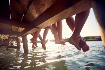 friends sitting on the dock and  enjoying together at holiday dangling legs in the water - 616158723