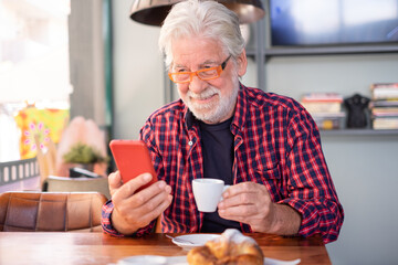 Smiling white-haired senior man sitting at cafe table holding an espresso coffee cup while looking at smart phone. Elderly caucasian male in eyeglasses enjoying breakfast - Powered by Adobe