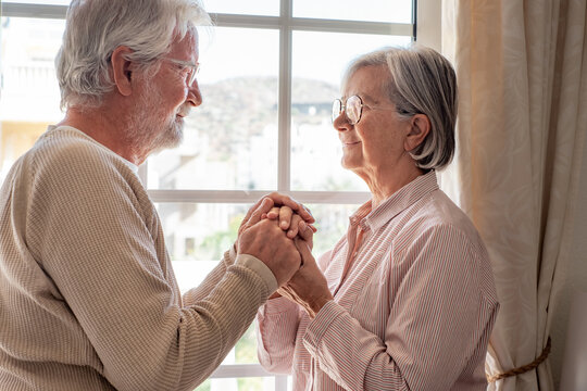 Senior family couple of Caucasian people looking in the eyes in front the window, elderly man and woman holding hands in a moment of melancholy