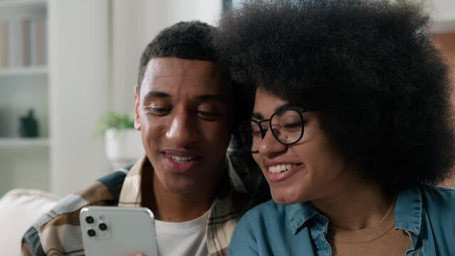 African American Couple Man And Woman Together At Home Looking At Phone Using Mobile Device Gadget To Search Offer Choosing Goods Online Booking Tickets For Vacations Discussing Talking Family Budget