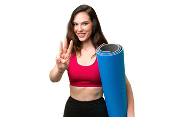 Young sport woman going to yoga classes while holding a mat over isolated background happy and counting three with fingers