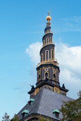 Fototapeta premium Copenhagen, Denmark - September 14, 2010: Vor Frelsers church. Tower closeup against blue sky. Golden trim of spiral and clock. Statues on corniche and green foliage peeps up to black roof