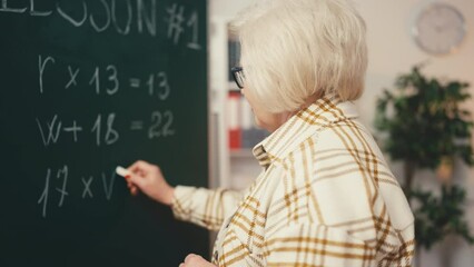 Senior woman math teacher writing equation on chalkboard, teaching a lesson