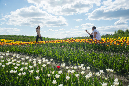 A guy takes a picture of his girlfriend with his phone at field of colorful tulips background.