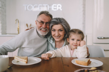 Smiling grandparents hugging granddaughter while having sandwiches