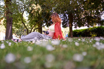 Young pregnant woman applying headphones on belly to listen to music in shady park on the summer day.regnancy and maternity concept.Pregnancy women is relaxing with music and headphone in outdoor.