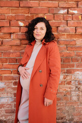 A happy pregnant woman in an orange coat against a city brick wall, touching her belly and looking at the camera